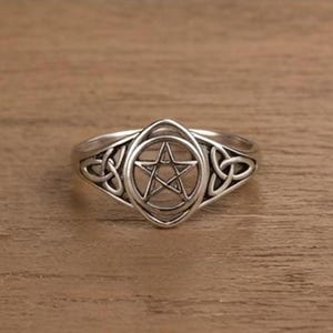 Peut inclure: Bague argentée ornée d'un pentagramme dans un motif de nœud celtique. La bague présente un centre ovale avec une étoile à cinq branches. La monture est ornée de nœuds complexes de chaque côté du motif central. La bague est posée sur une surface en bois.