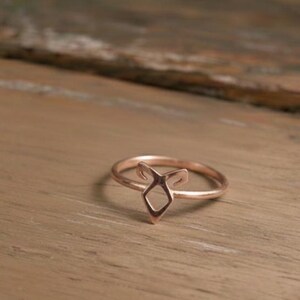 K&ouml;nnte beinhalten: Ein ros&eacute;goldener Ring mit geometrischem Design. Der Ring hat eine zentrale Diamantenform mit zwei nach oben zeigenden Elementen, die einen stilisierten Fuchskopf bilden. Der Ring ist vor einem Holzgrund platziert.