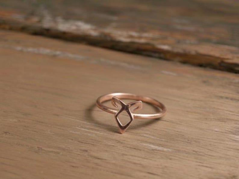 K&ouml;nnte beinhalten: Ein ros&eacute;goldener Ring mit geometrischem Design. Der Ring hat eine zentrale Diamantenform mit zwei nach oben zeigenden Elementen, die einen stilisierten Fuchskopf bilden. Der Ring ist vor einem Holzgrund platziert.