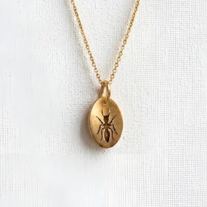 May include: A gold-colored pendant necklace featuring an oval pendant with a cut-out ant design. The ant is centered within the oval, and the necklace chain is delicate. The background is a textured white surface.