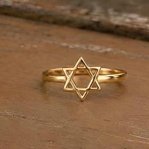 May include: A gold-colored ring featuring a Star of David design. The ring is set against a wooden surface, showcasing the intricate details of the six-pointed star. The ring's band is smooth and polished.