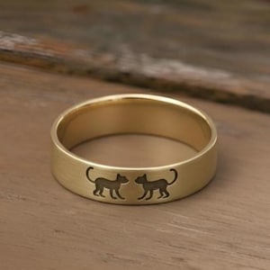May include: A gold-coloured ring with a matte finish. The ring features two black cat silhouettes engraved around the band. The ring is displayed on a wooden surface.