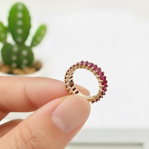 Peut inclure: Une bague dorée ornée d'une rangée continue de pierres précieuses rectangulaires rouge foncé. La bague est présentée sur un fond blanc, mettant en valeur la couleur vive des pierres. Le design de la bague est simple, mais élégant, adapté à un usage quotidien ou à des occasions spéciales.