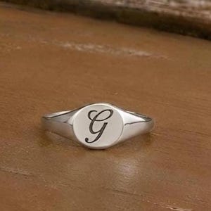 Könnte beinhalten: Ein silberner Siegelring mit poliertem Finish. Der Ring hat eine kreisförmige Oberfläche mit dem Buchstaben "G" in schwarzer, kursiver Schrift. Der Ring liegt auf einer braunen Holzoberfläche.