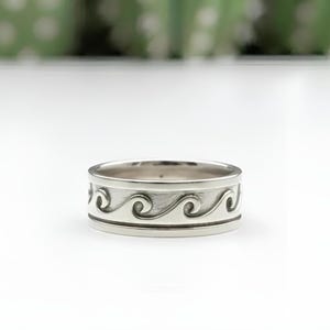 Peut inclure: Une bague en argent ornée d'un motif de vagues. La bague présente un anneau orné d'une série de vagues stylisées, créant un motif continu sur tout le pourtour. La bague est posée sur un fond blanc.