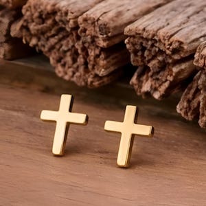 Op de afbeelding: Twee goudkleurige kruisvormige oorstekers. De oorbellen worden gepresenteerd op een houten oppervlak, met stukken getextureerd hout op de achtergrond. De kruisen hebben een eenvoudig, klassiek ontwerp.