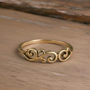 May include: A gold-colored ring with an ornate, swirling design. The ring features three decorative sections with a continuous, flowing pattern. The ring is set against a light brown wooden surface.