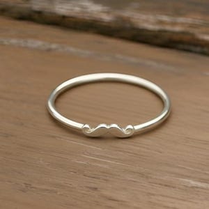 May include: A silver ring with a mustache design. The ring is thin and circular, with a small mustache charm centered on the band. The ring is set against a wooden surface.