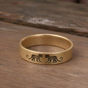 Könnte beinhalten: Ein goldfarbener Ring mit mattem Finish. Der Ring zeigt zwei geätzte Affensilhouetten, die sich gegenüberstehen. Der Ring ist etwa 0,8 cm breit und wird auf einer Holzoberfläche präsentiert.