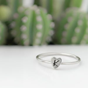 Puede incluir: Un anillo plateado con un dije en forma de corazón con la letra "K" en negro. El anillo se encuentra sobre un fondo borroso de plantas verdes. Es una joya delicada.