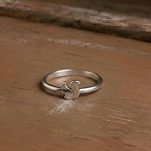 May include: A silver ring featuring a squirrel silhouette. The ring is set against a wooden surface, highlighting the ring's design. The squirrel is a simple, yet elegant, design.