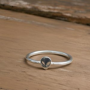Può includere: Un anello d'argento con un design a testa di alieno. L'anello ha una fascia sottile e una piccola faccia aliena ovale come elemento centrale. L'anello è esposto su una superficie di legno.