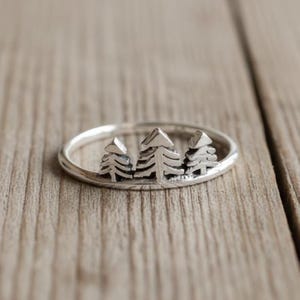 May include: A silver ring featuring a forest scene with three detailed pine trees. The ring has a simple band and the trees are the focal point of the design. The ring is shown on a wooden surface.