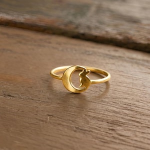 Puede incluir: Un anillo dorado con un diseño de media luna y una silueta de gato. El anillo se encuentra sobre un fondo de madera, resaltando los detalles de la joya. El anillo es una pieza de joyería.