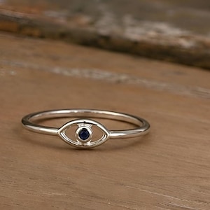 Puede incluir: Anillo de plata con diseño de ojo y una pequeña piedra redonda azul oscuro en el centro. El anillo se muestra sobre una superficie de madera, resaltando sus delicados detalles y artesanía.