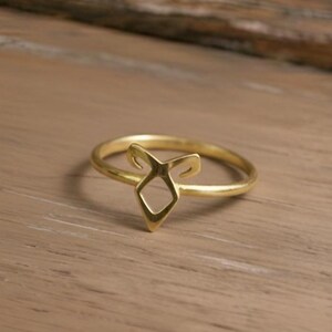 K&ouml;nnte beinhalten: Ein goldfarbener Ring mit einem einzigartigen, offenen Design. Der Ring hat eine zentrale Diamantenform mit stilisierten H&ouml;rnern oder Kurven, die sich nach oben erstrecken. Der Ring liegt auf einer hellbraunen Holzoberfl&auml;che.