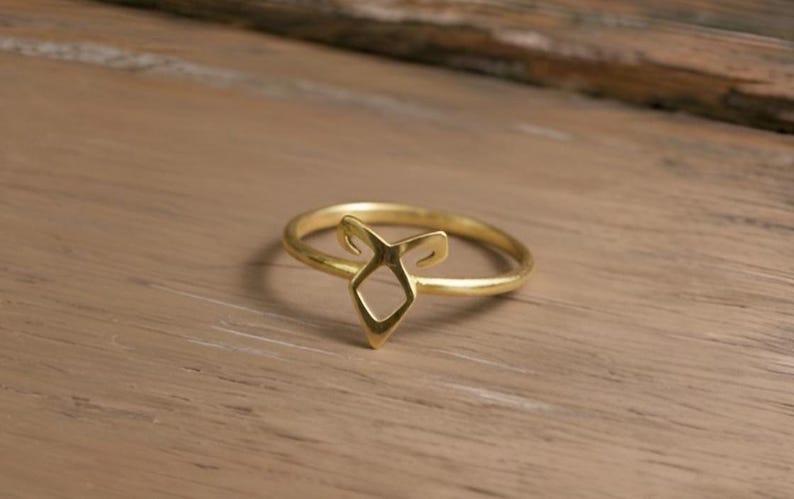 K&ouml;nnte beinhalten: Ein goldfarbener Ring mit einem einzigartigen, offenen Design. Der Ring hat eine zentrale Diamantenform mit stilisierten H&ouml;rnern oder Kurven, die sich nach oben erstrecken. Der Ring liegt auf einer hellbraunen Holzoberfl&auml;che.