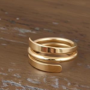 May include: A gold-coloured spiral ring with a smooth, polished finish. The ring wraps around the finger in a continuous, flowing design. The ring is made of a shiny, reflective metal.