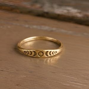 May include: A gold-toned ring featuring a series of engraved moon phase symbols along the front band. The design includes a central full circle flanked by crescent shapes that gradually decrease in size, creating a continuous lunar cycle pattern.