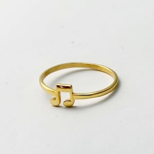 May include: A gold-colored ring featuring a musical eighth note design. The ring is a simple band with the note as a prominent, openwork element. The ring is set against a white background.