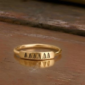 Puede incluir: Anillo dorado con una cara rectangular con un diseño grabado de seis pinos estilizados. El anillo se encuentra sobre un fondo de madera borroso, resaltando los detalles de la joya.