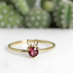 Puede incluir: Un anillo dorado con un diseño de conejito. El anillo presenta una gema redonda de color rosa intenso para la cara del conejito y dos pequeños bucles curvos para las orejas. El anillo está sobre un fondo borroso verde y blanco.