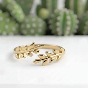 May include: A gold-coloured ring with a leaf design. The ring features two leaf branches that wrap around the finger. The ring is set against a white surface with a blurred background of green plants.