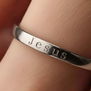 May include: A close-up of a silver ring with the word "Jesus" engraved on the band. The ring is worn on a finger, and the metal has a polished, reflective surface. The ring is a religious jewellery item.