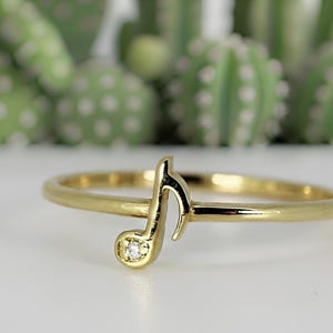 May include: A gold-colored ring featuring a musical note design with a small, clear gemstone. The ring is set against a blurred background of green and white, suggesting a nature-inspired theme.
