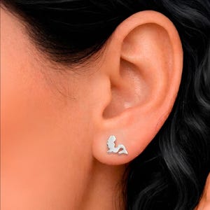 May include: Silver mermaid stud earrings. The earrings are small and feature a mermaid silhouette. The earrings are shown on an ear, with black hair in the background. The earrings are a simple, elegant design.