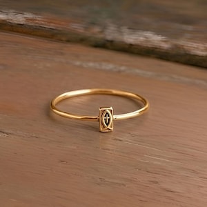 Könnte beinhalten: Ein goldfarbener Ring mit einem rechteckigen Mittelstück mit einem Kreuzdesign. Der Ring ist dünn und zart, mit poliertem Finish. Der Ring liegt auf einer Holzoberfläche.