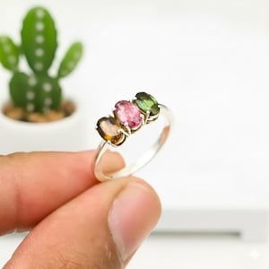 Op de afbeelding: Een zilveren ring met drie ovale edelstenen in bruin, roze en groen. De edelstenen zijn in een rij geplaatst, wat een kleurrijk en elegant ontwerp creëert. De ring wordt tegen een witte achtergrond getoond.