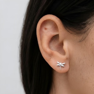 Peut inclure: Une petite boucle d'oreille à clou de couleur argentée polie en forme de libellule. Le bijou présente un corps fin et quatre ailes délicates et symétriques, fixé solidement au lobe de l'oreille par un fermoir à tige pour un aspect épuré.