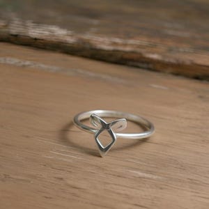K&ouml;nnte beinhalten: Ein silberner Ring mit geometrischem Design. Der Ring hat eine zentrale Diamantenform mit zwei nach oben gerichteten, gebogenen Elementen. Der Ring liegt auf einer Holzoberfl&auml;che.