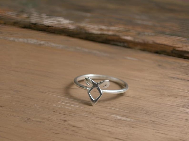K&ouml;nnte beinhalten: Ein silberner Ring mit geometrischem Design. Der Ring hat eine zentrale Diamantenform mit zwei nach oben gerichteten, gebogenen Elementen. Der Ring liegt auf einer Holzoberfl&auml;che.