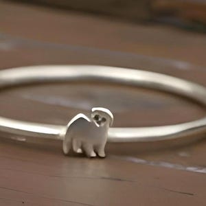 May include: A silver bracelet featuring a small llama charm. The circular bracelet is displayed on a brown wooden surface. The llama charm is positioned centrally on the bracelet.