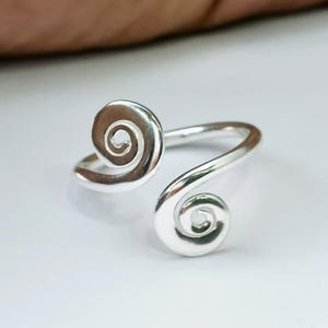 Könnte beinhalten: Ein silberfarbener Ring mit einem Spiraldesign an jedem Ende. Der Ring hat eine glatte, polierte Oberfläche und ist vor einem weißen Hintergrund platziert. Das Spiraldesign ist ein herausragendes Merkmal des Rings.