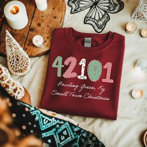 May include: A maroon sweatshirt with the number "42101" appliqued in green gingham, red gingham, and white fabric with a holly pattern. The text "Bowling Green, Ky Small Town Christmas" is below the numbers.