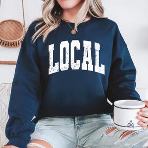 May include: Navy blue sweatshirt with the word "LOCAL" in distressed white block letters. The sweatshirt is long-sleeved and has a crew neck. The person is wearing light blue denim shorts and holding a white mug.