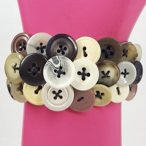 May include: A bracelet made of a variety of buttons in different colors and patterns, including brown, white, and black. The buttons are arranged in a circular pattern and are attached to a clear elastic band.