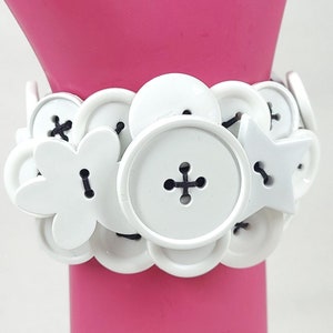 May include: White button bracelet made of various sized and shaped buttons. The bracelet is made of white plastic buttons with a black thread running through the holes of the buttons.