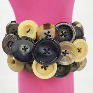 May include: A button bracelet featuring a variety of colors and sizes. The bracelet is made of multiple buttons in shades of black, brown, beige, and olive green. The buttons are attached together to form a bracelet.