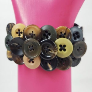 May include: A bracelet made of various buttons in black, brown, beige, and green colors. The buttons are sewn together with black thread.
