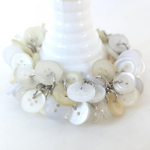 May include: A cluster of white and off-white buttons, each attached with a silver-colored ring, encircles the base of a white, cylindrical object. The buttons vary in size and shade, creating a textured, monochromatic design.