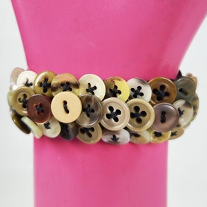 May include: A bracelet made of various brown, beige, and cream colored buttons sewn together with black thread.