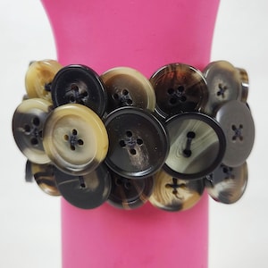 May include: A bracelet made of a cluster of brown and cream buttons with black stitching. The buttons are arranged in a circular pattern and are held together by a thin black cord.