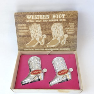 May include: A vintage Western Boot metal salt and pepper set in a box. The silver-colored boots have intricate detailing and a red "San Diego" emblem. The box is brown with text that reads "Western Boot Metal Salt and Pepper Sets."