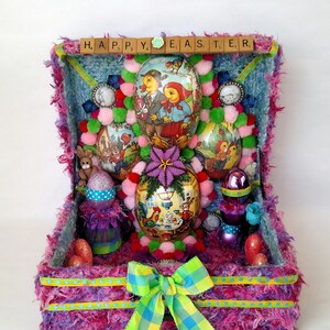 May include: A pink and purple fuzzy box with a yellow and green plaid bow. The box is open and filled with colorful Easter eggs, including a few with painted chicks and bunnies. The top of the box has a blue background with a row of wooden letter tiles spelling "HAPPY EASTER".