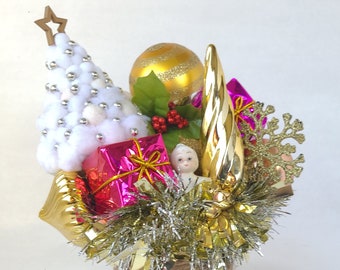 May include: A festive Christmas centerpiece featuring a white cotton tree with silver beads, a gold ornament, and wrapped pink gifts. The arrangement includes gold tinsel, a gold snowflake, and a small doll, all set in a twine-wrapped base.