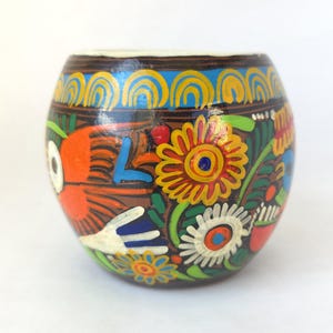 May include: A hand-painted, round, brown ceramic vessel with a white rim. The vessel is decorated with colorful floral and bird designs in orange, yellow, blue, and green. The vessel is likely used for decorative purposes.